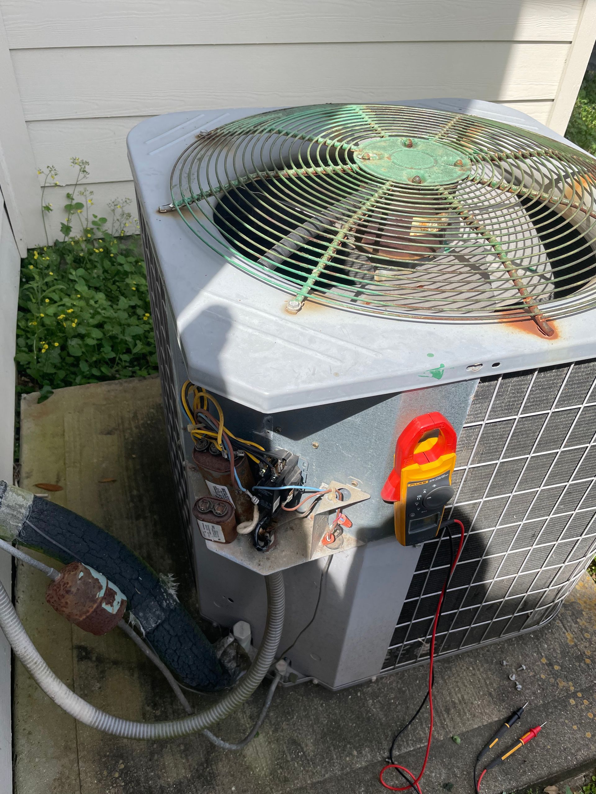An air conditioner unit with electrical wires exposed and a multimeter attached outdoors.