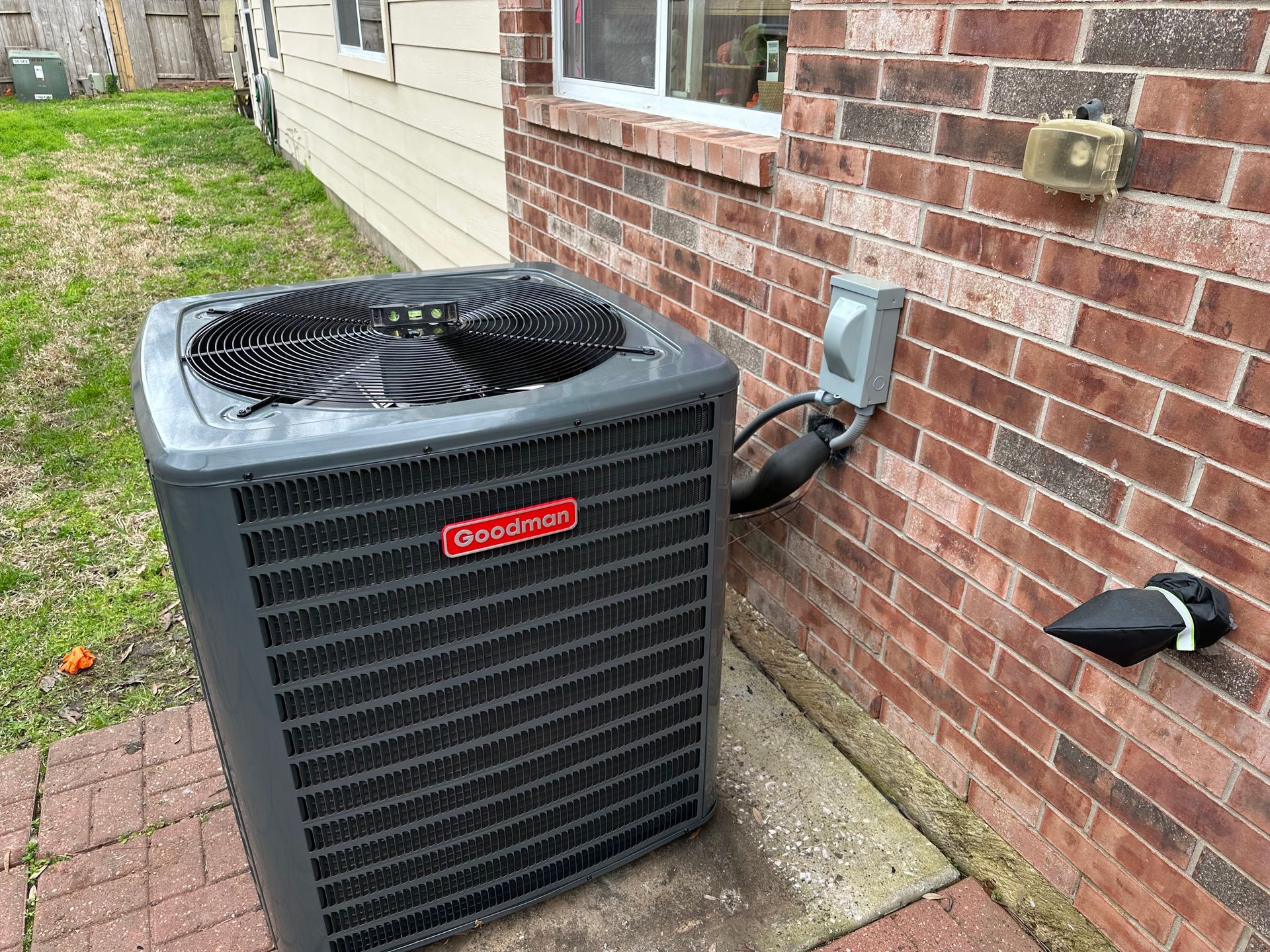 Gray Goodman AC unit next to a brick building. Electrical box and water spigot on the wall.