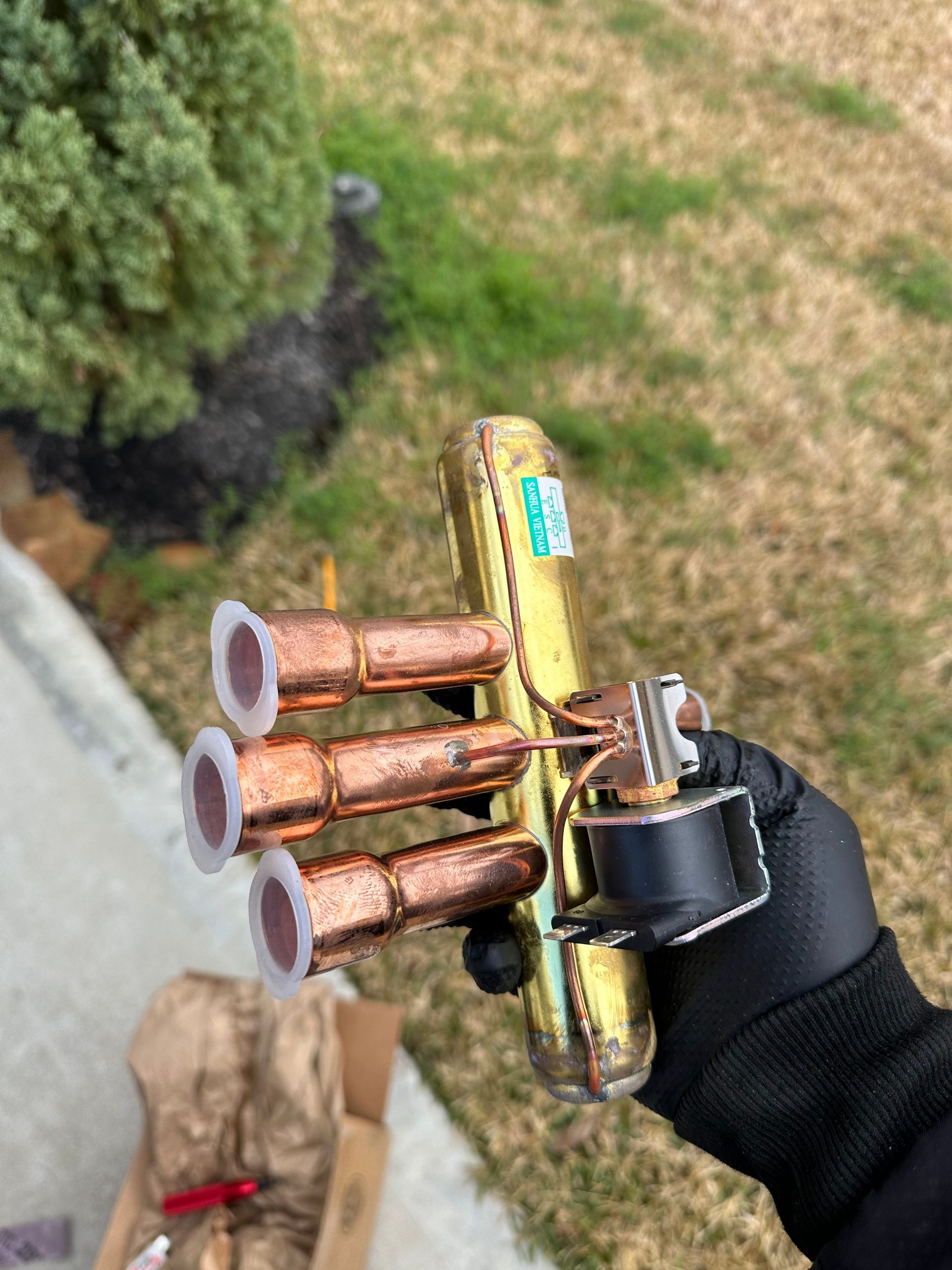 Hand holding a copper HVAC valve with multiple ports and a solenoid, outdoors near greenery.