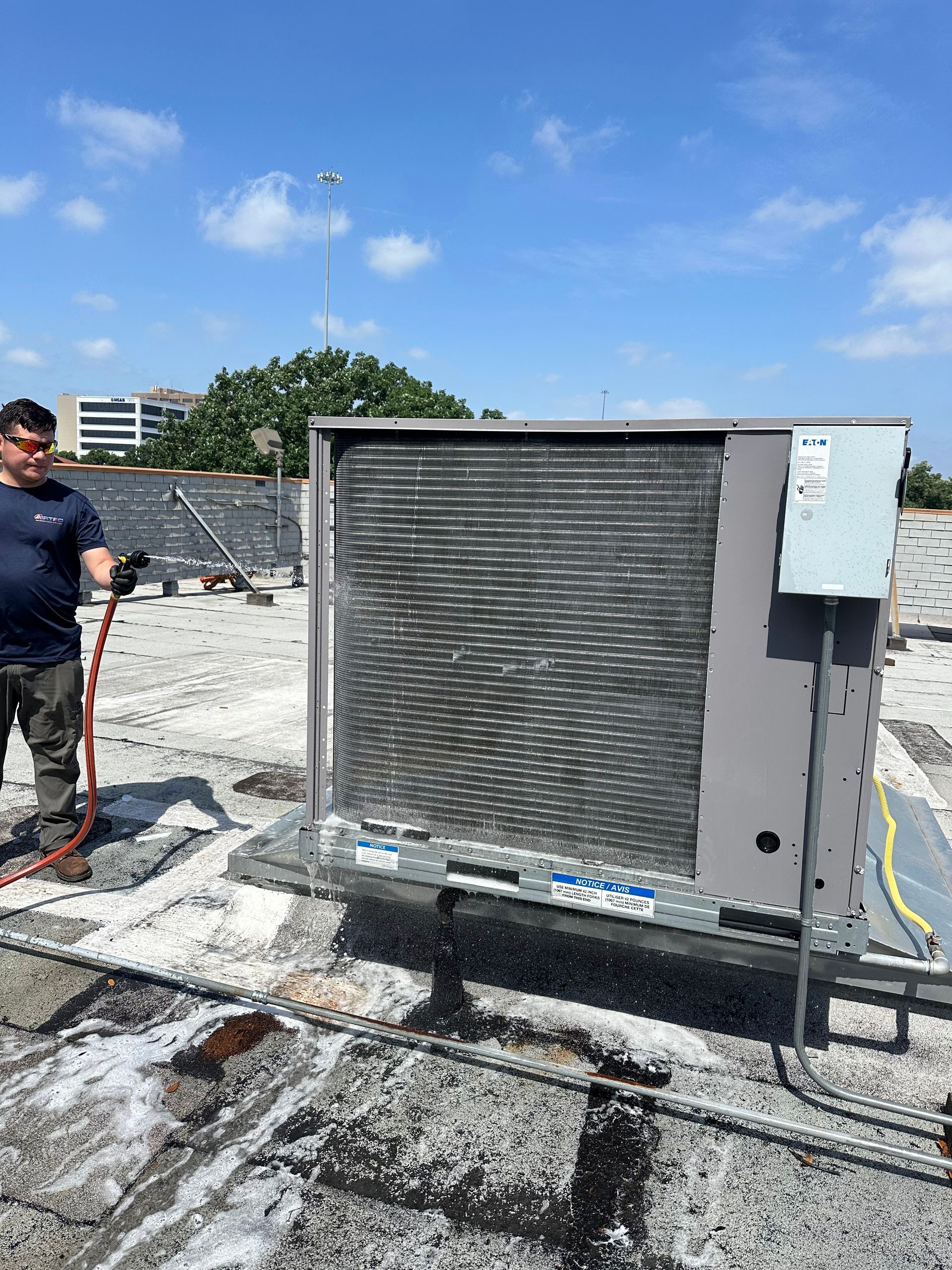 Person sprays water on an AC unit on a rooftop under a blue sky.