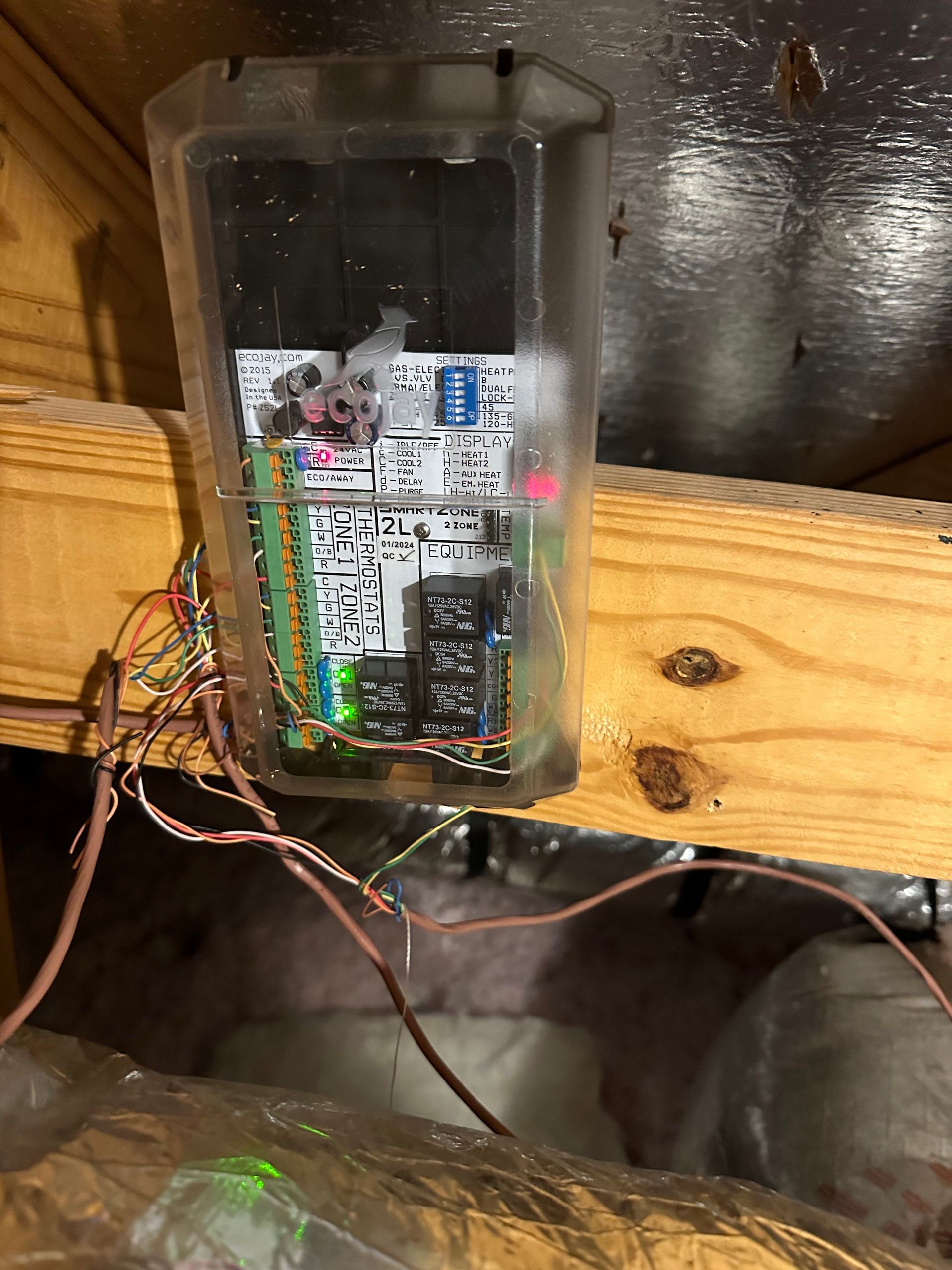 Electronic circuit board inside a clear plastic enclosure mounted on a wooden beam, wires extending downwards.
