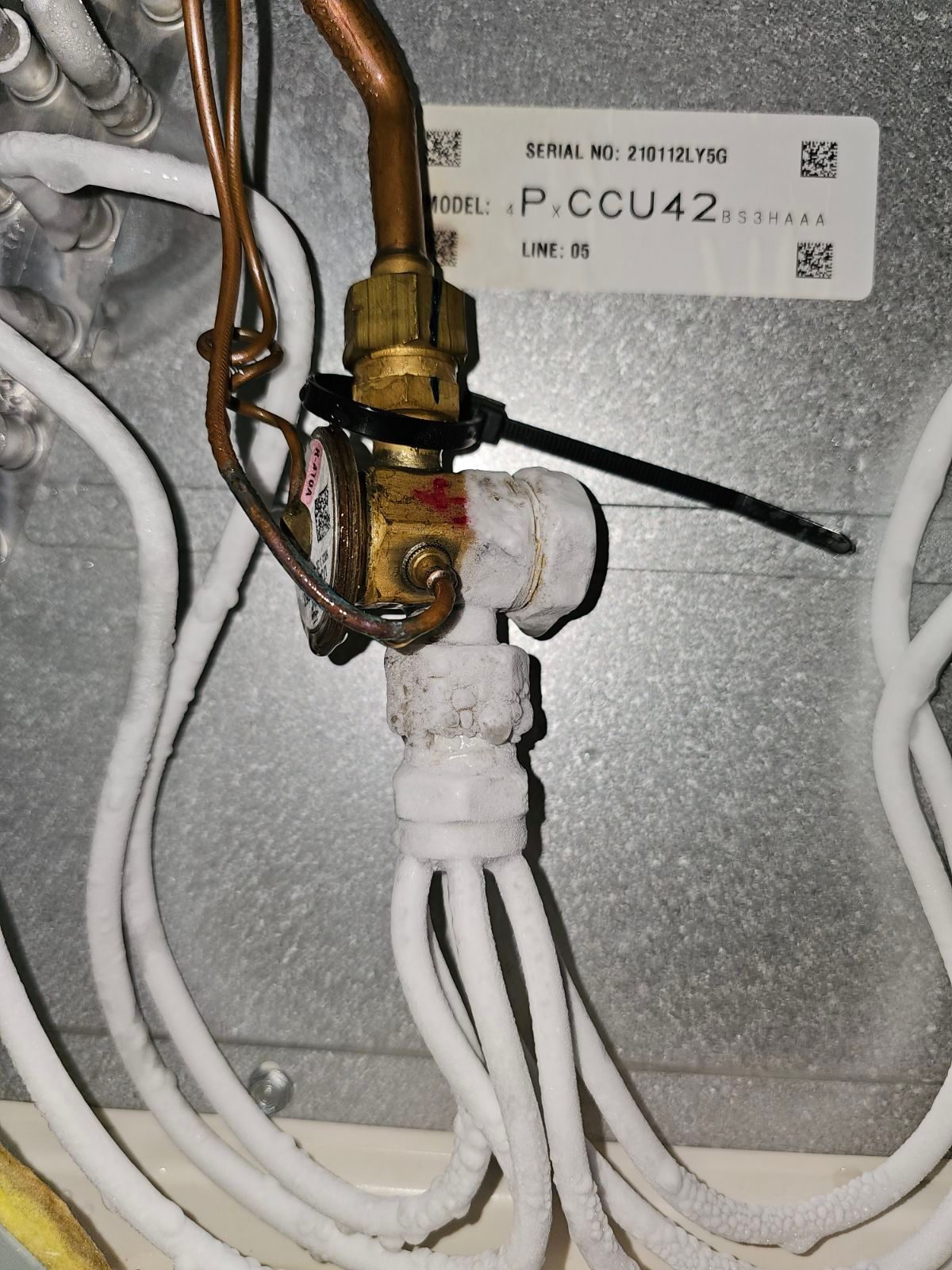 Close-up of a frozen air conditioning valve with copper tubing, white insulation, and a serial number label.