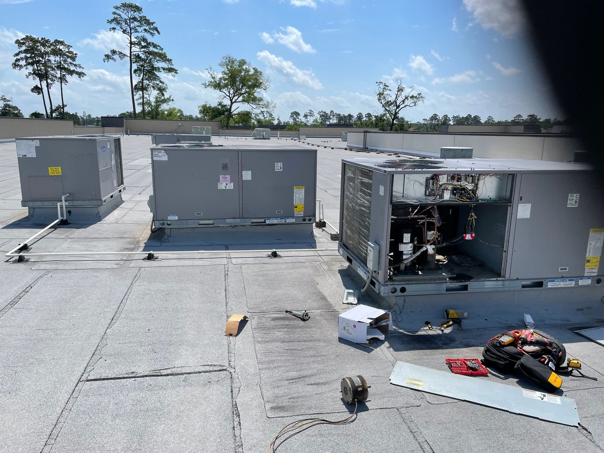 Rooftop view of three HVAC units on a flat roof, one unit open for maintenance with tools and components visible.