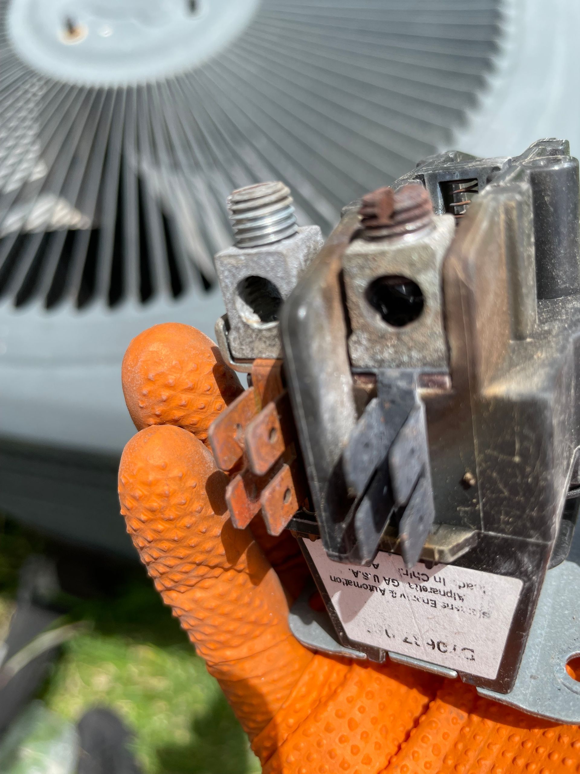 Hand holding an electrical component with screw terminals, next to an air conditioner unit.
