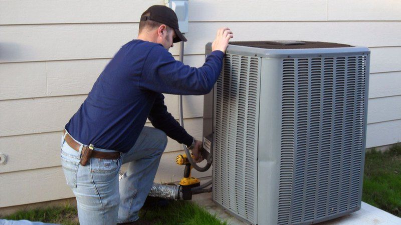 Man kneeling, working on outdoor air conditioner unit, using a drill.