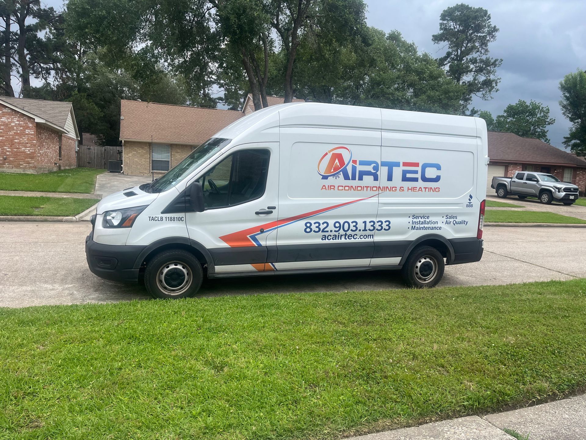 White AIRTEC service van parked on a residential street; trees and houses in the background.