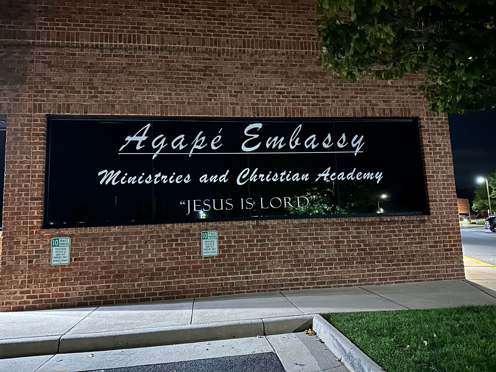 Brick church sign reading “Agape Embassy Ministries and Christian Academy” at night, with “Jesus is Lord” below.