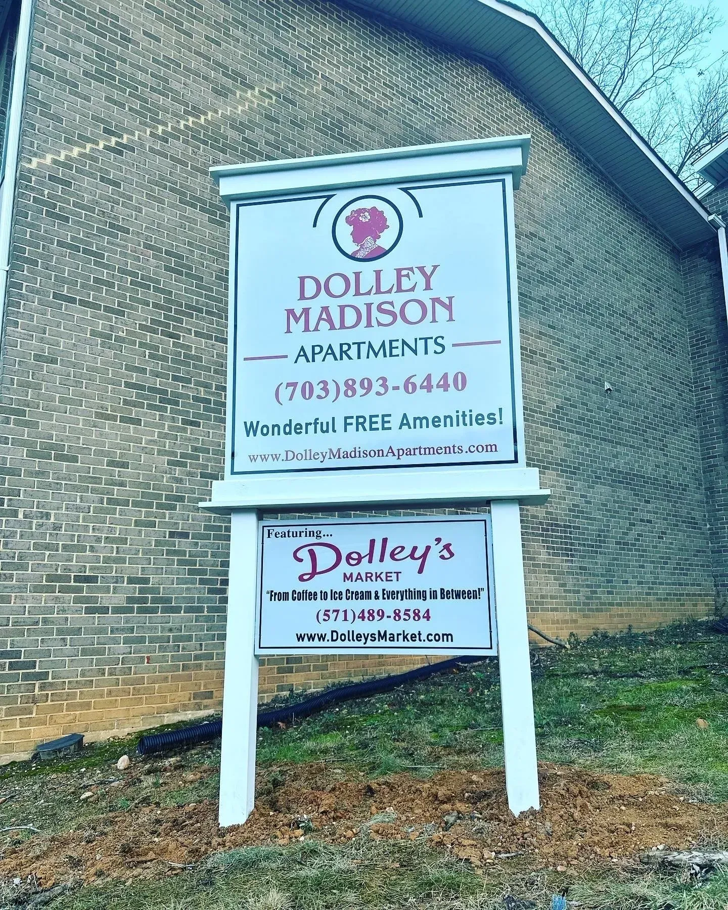 A dual-panel sign for Dolley Madison Apartments and Dolley's Market, set against a brick building on a grassy slope.