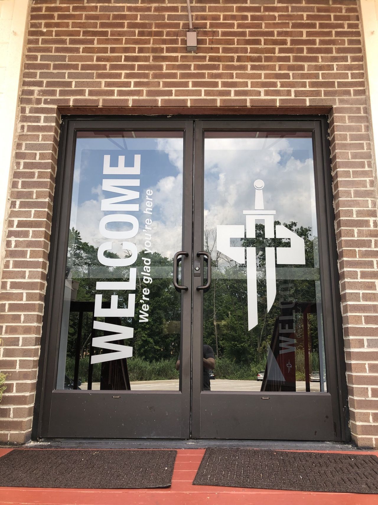 Glass church doors with a large white “WELCOME” sign and a cross symbol reflected on the panels