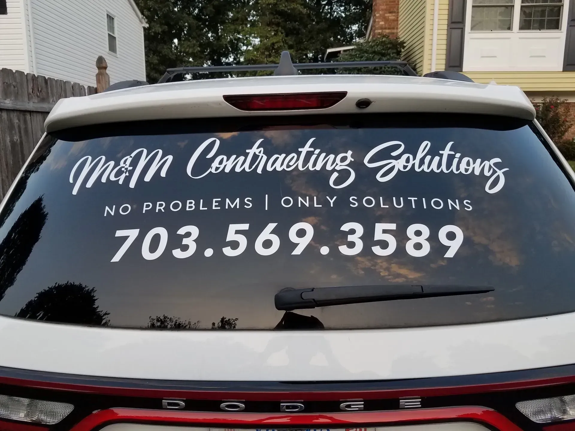 Back window of a white Dodge vehicle displaying white vinyl lettering for M&M Contracting Solutions, phone 703-569-3589.