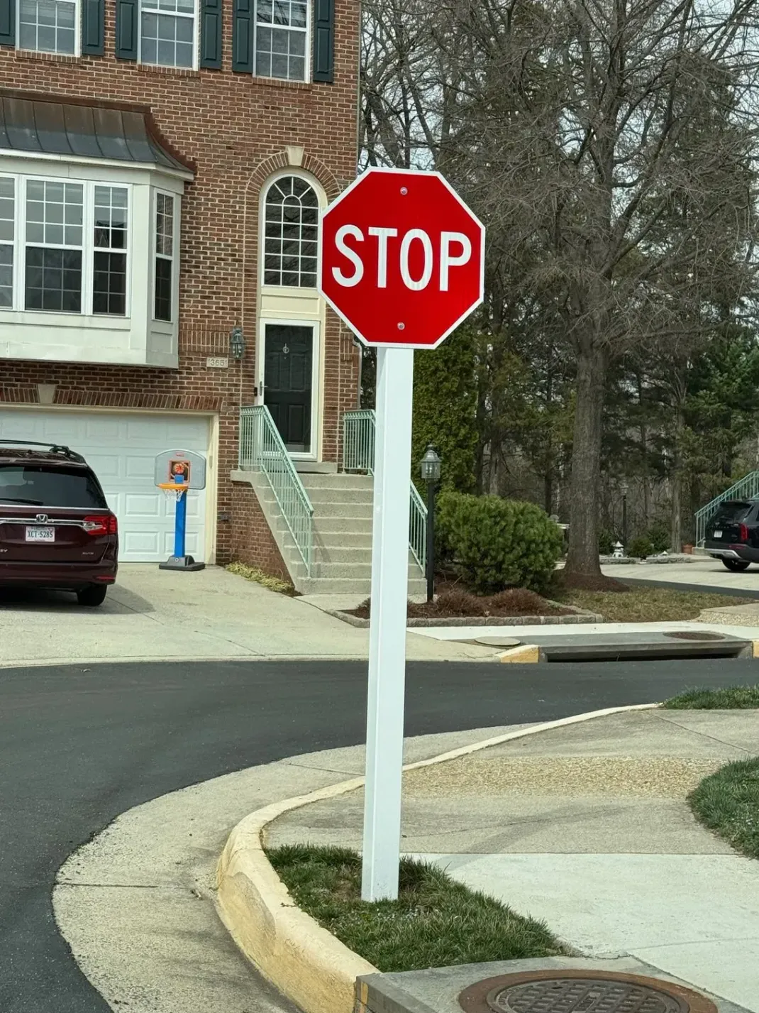 A red, octagonal stop sign mounted on a white pole in front of a brick suburban house and a parked car.