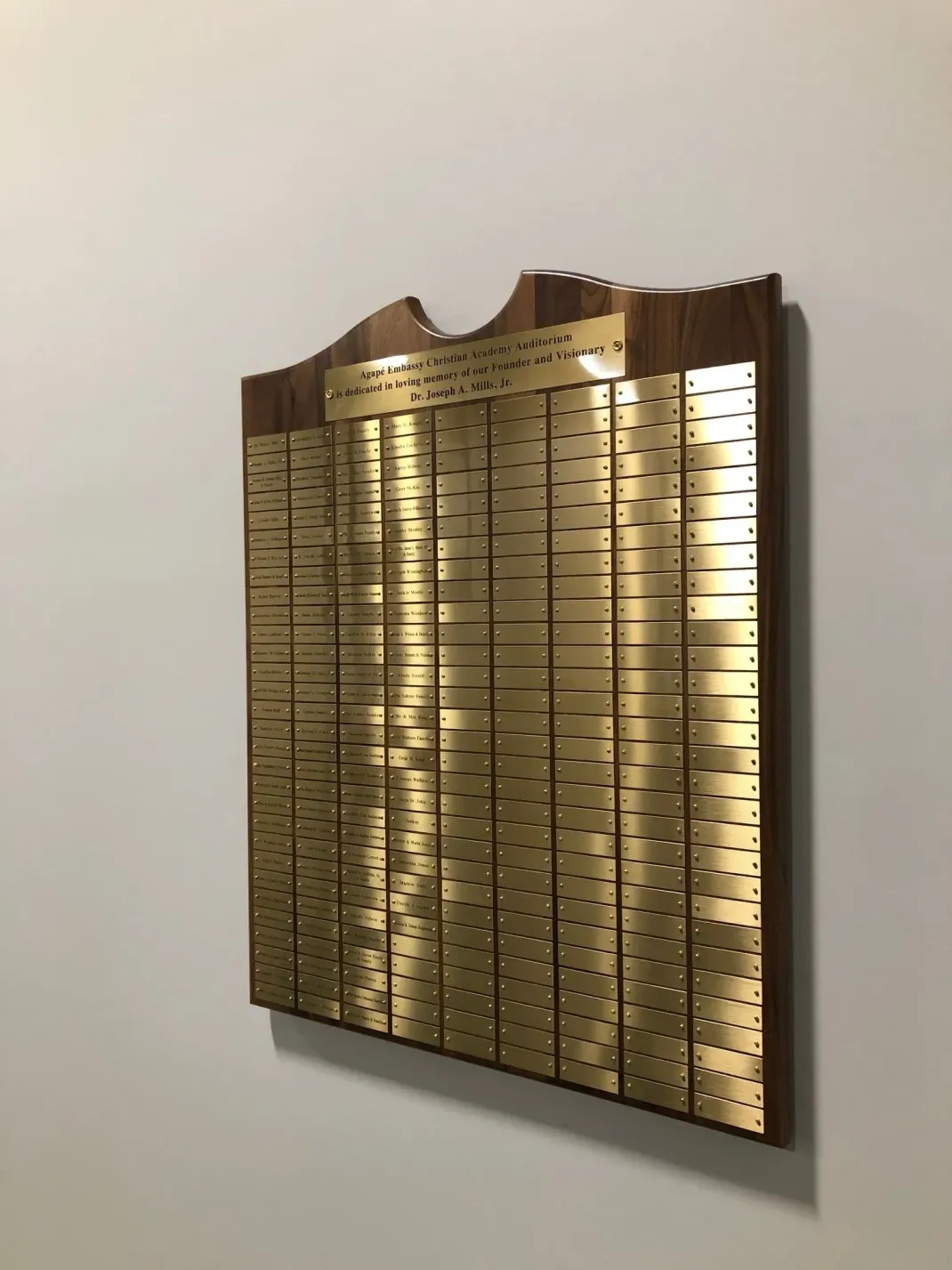 A wall-mounted wooden recognition plaque featuring many small, gold-colored metal nameplates arranged in columns.