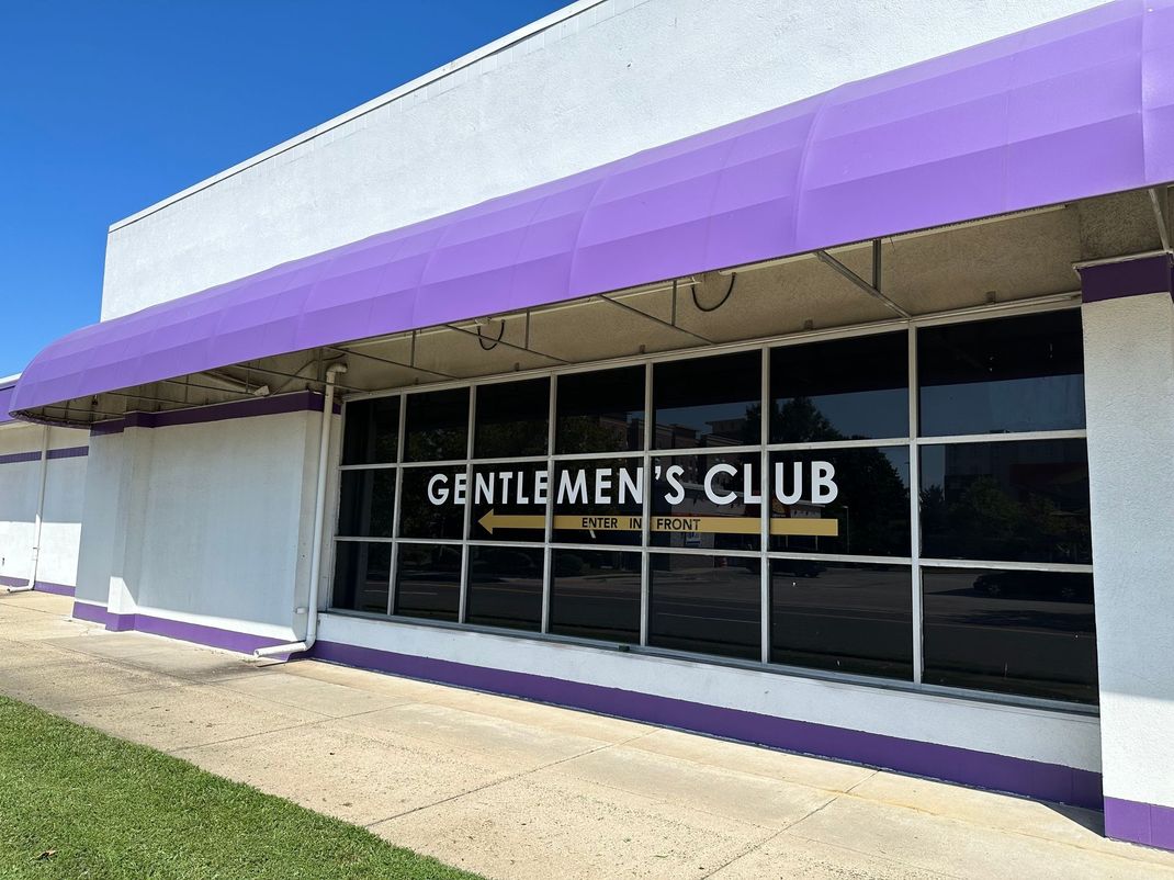 Storefront with purple awning and “Gentlemen’s Club” sign on a sunny street.