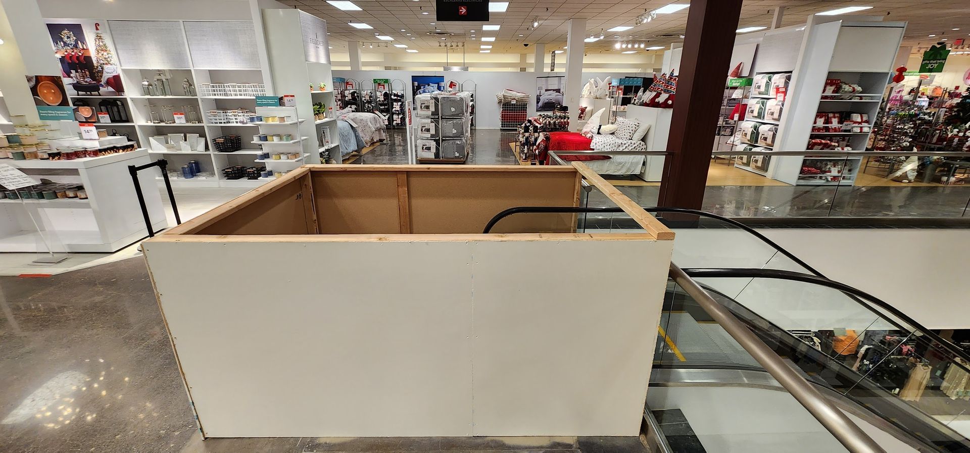 A wooden box is sitting in the middle of a shopping mall next to an escalator.