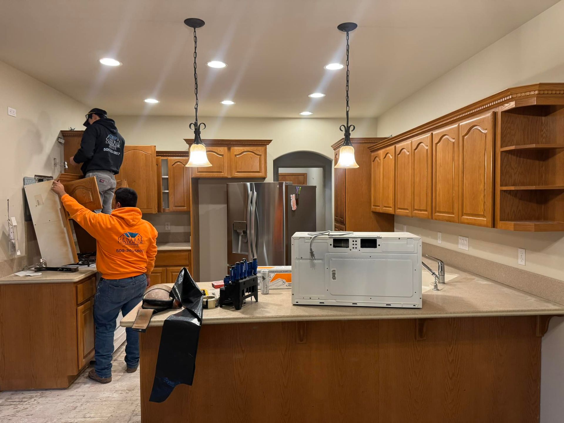 Two men are working in a kitchen with wooden cabinets.