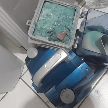 A blue vacuum cleaner is sitting on a tiled floor next to a broken glass container.