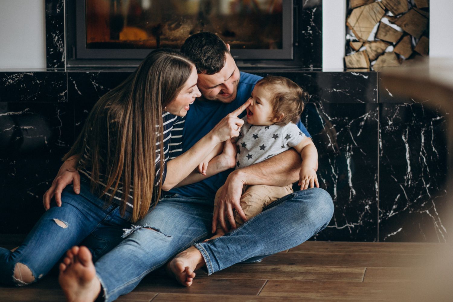 Family of three sitting on floor, interacting with a baby; near fireplace.