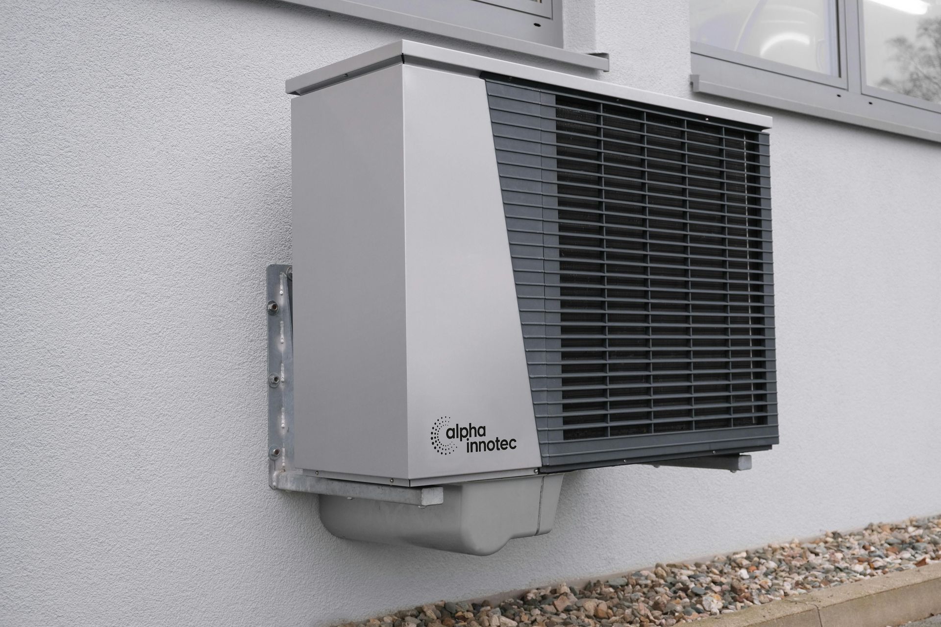 Exterior heat pump mounted on a white wall. Gray unit with a black grate, near a window and gravel.