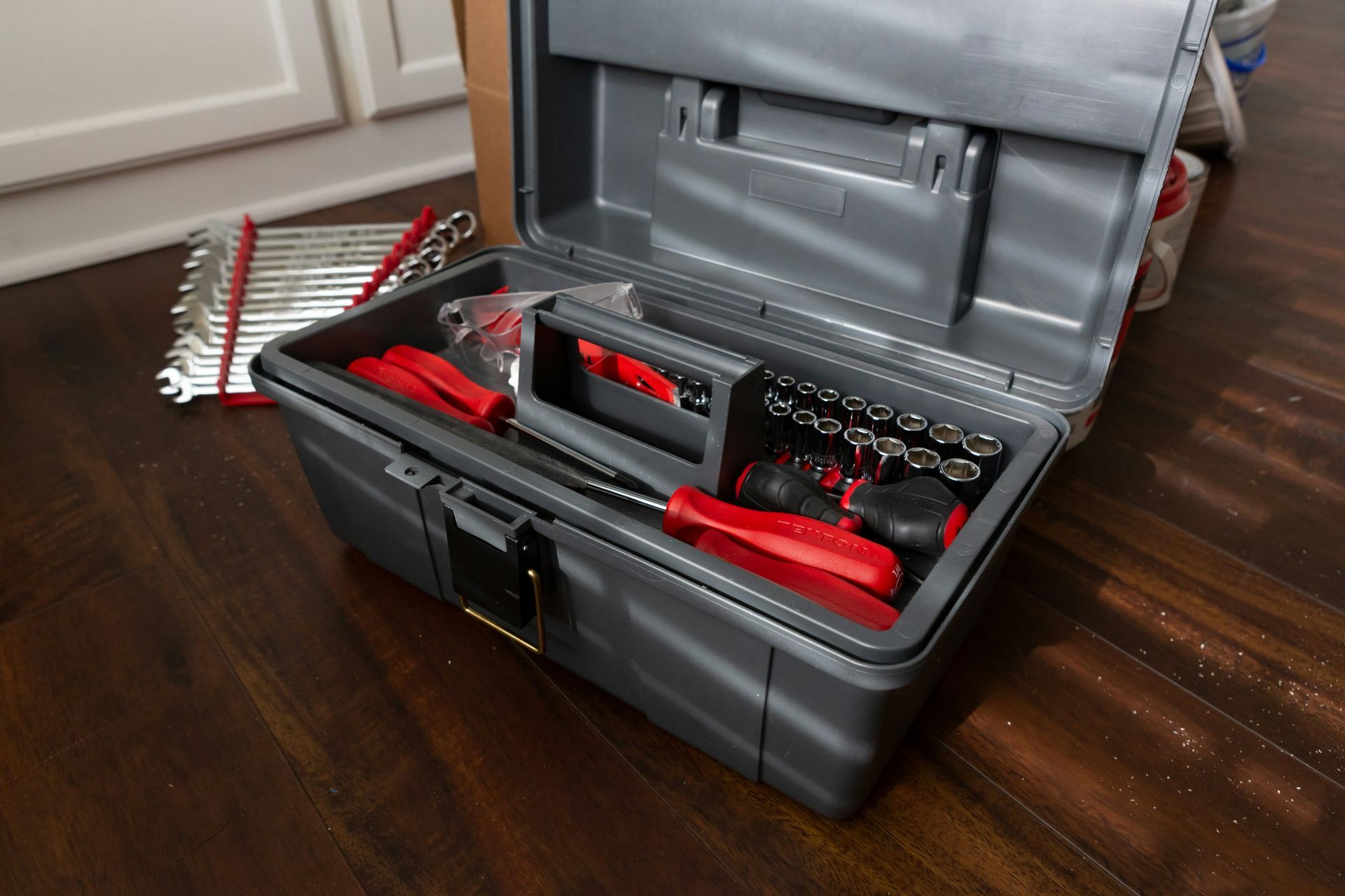 Open gray toolbox with red-handled tools and sockets on a wood floor.