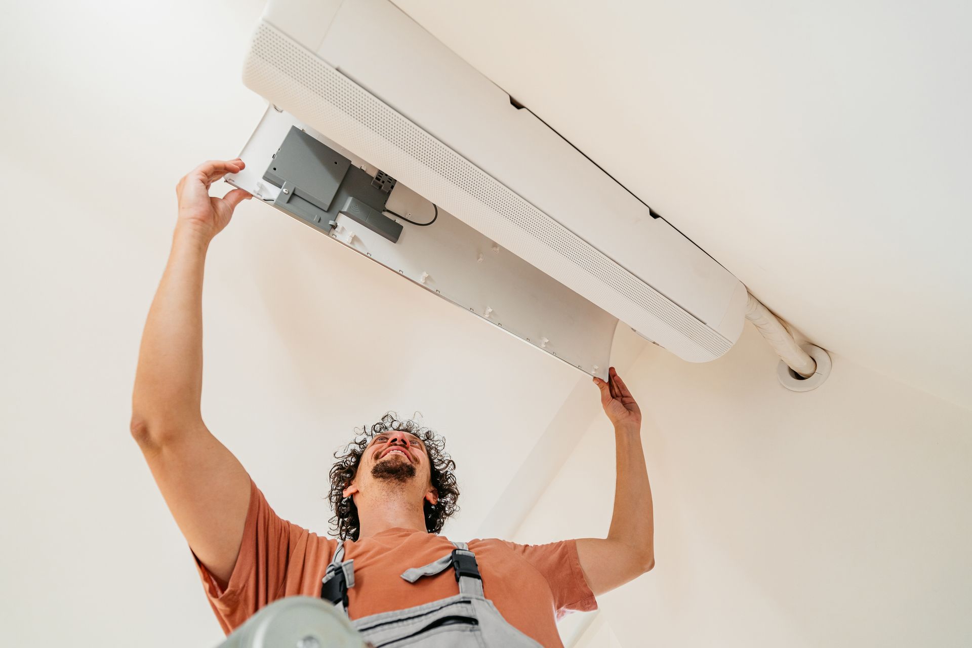 Man on a ladder cleaning a white air conditioner on a white wall.