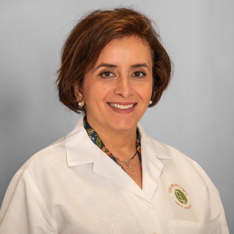 Woman in a white coat smiles at the camera; she has short brown hair and a patterned shirt.