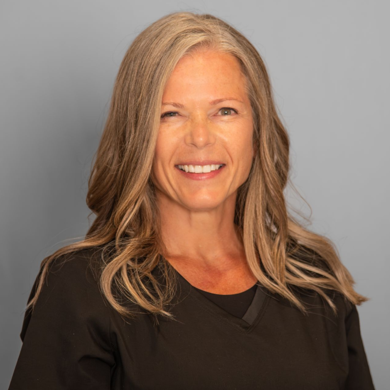 Woman with long gray hair smiles, wearing a black top against a gray background.