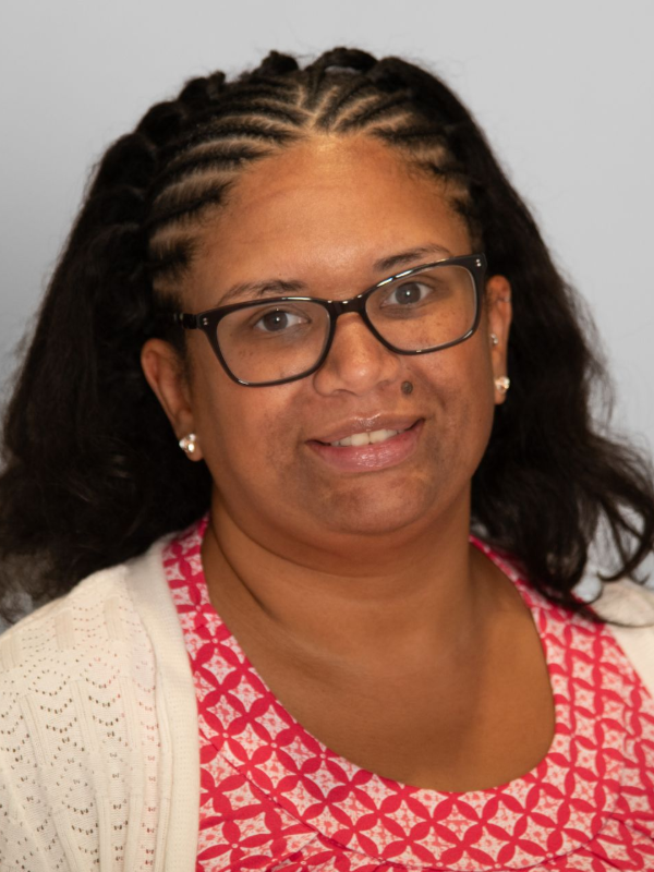 Woman with dark hair in braids and glasses smiles, wearing a red patterned shirt and a cream cardigan.