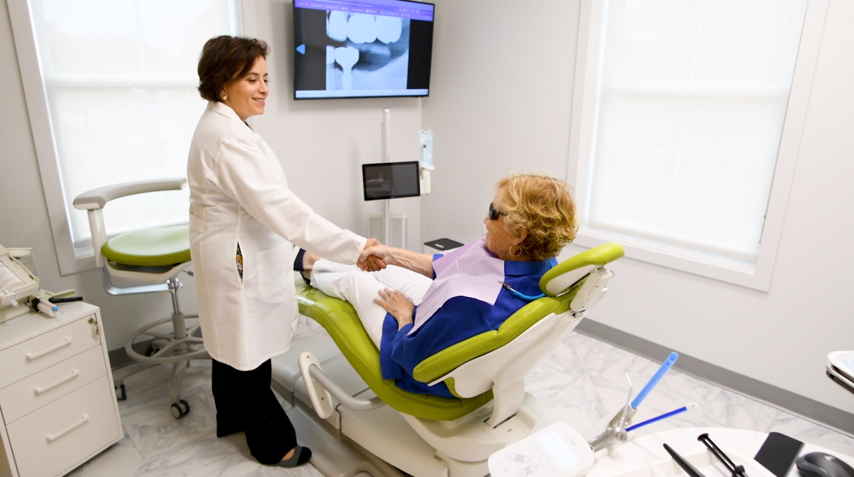 Woman in dental chair, holding jaw, appearing to be in pain; dental assistant in background.