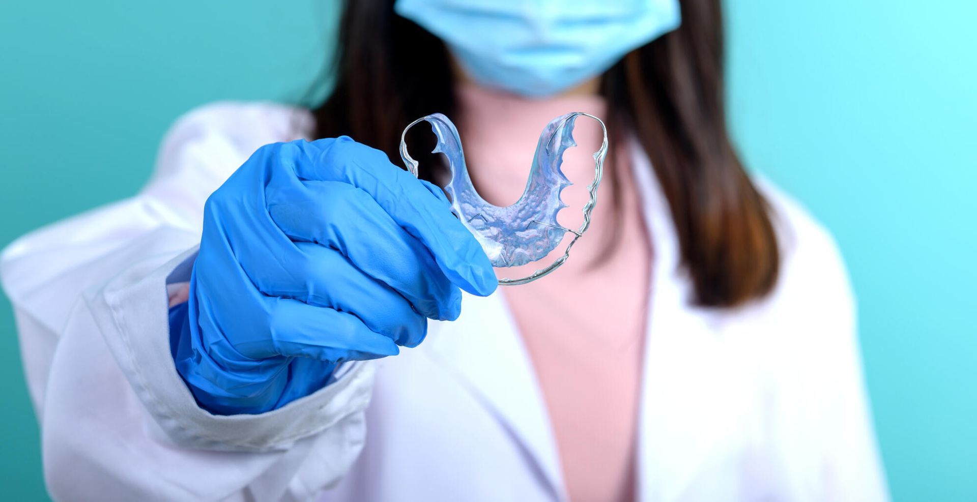 A person in a white lab coat and blue face mask holds up a clear dental retainer while wearing blue medical gloves.