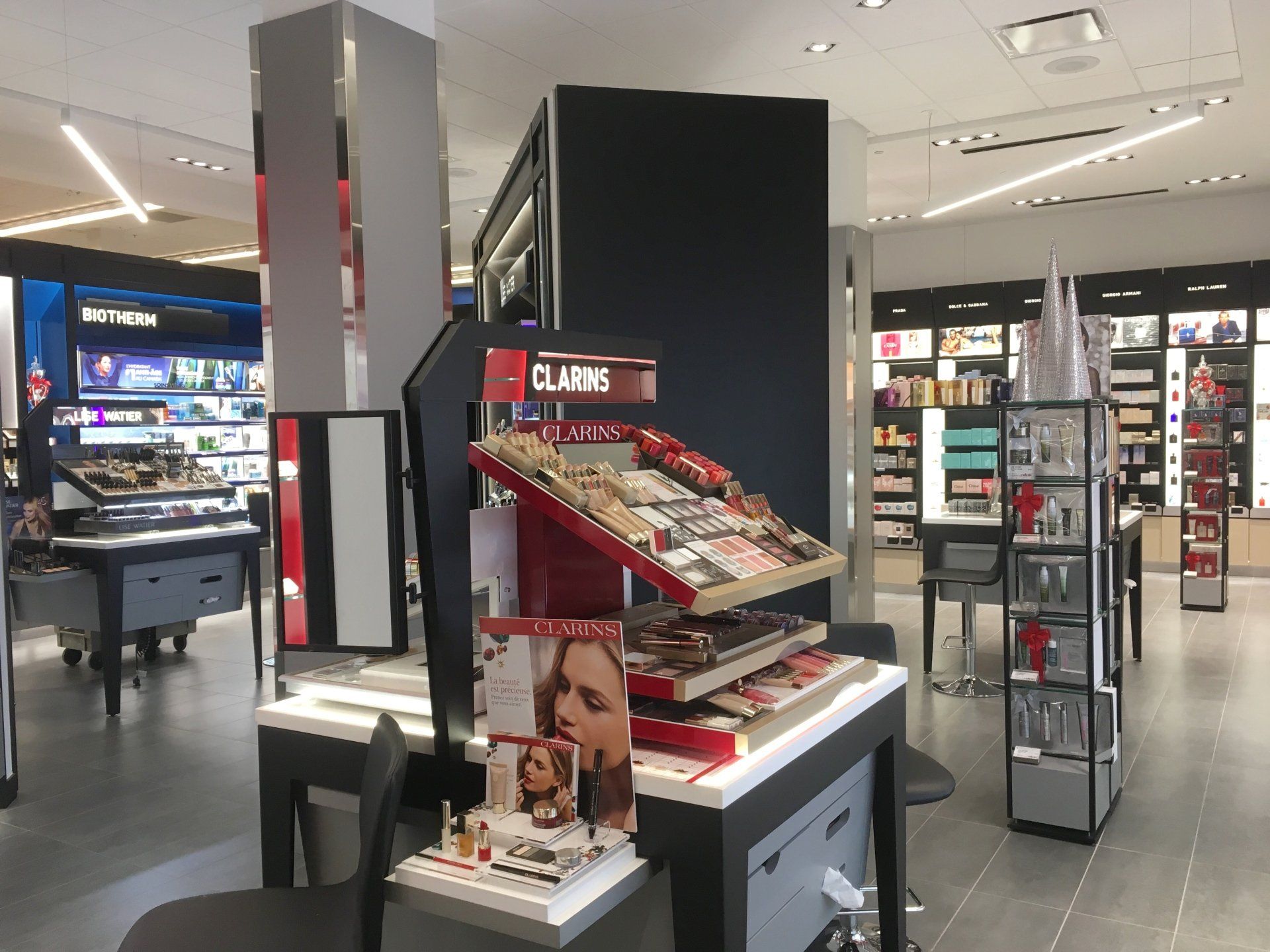 Présentoir de maquillage Clarins dans un magasin lumineux. Des présentoirs rouges et noirs mettent en valeur les produits.