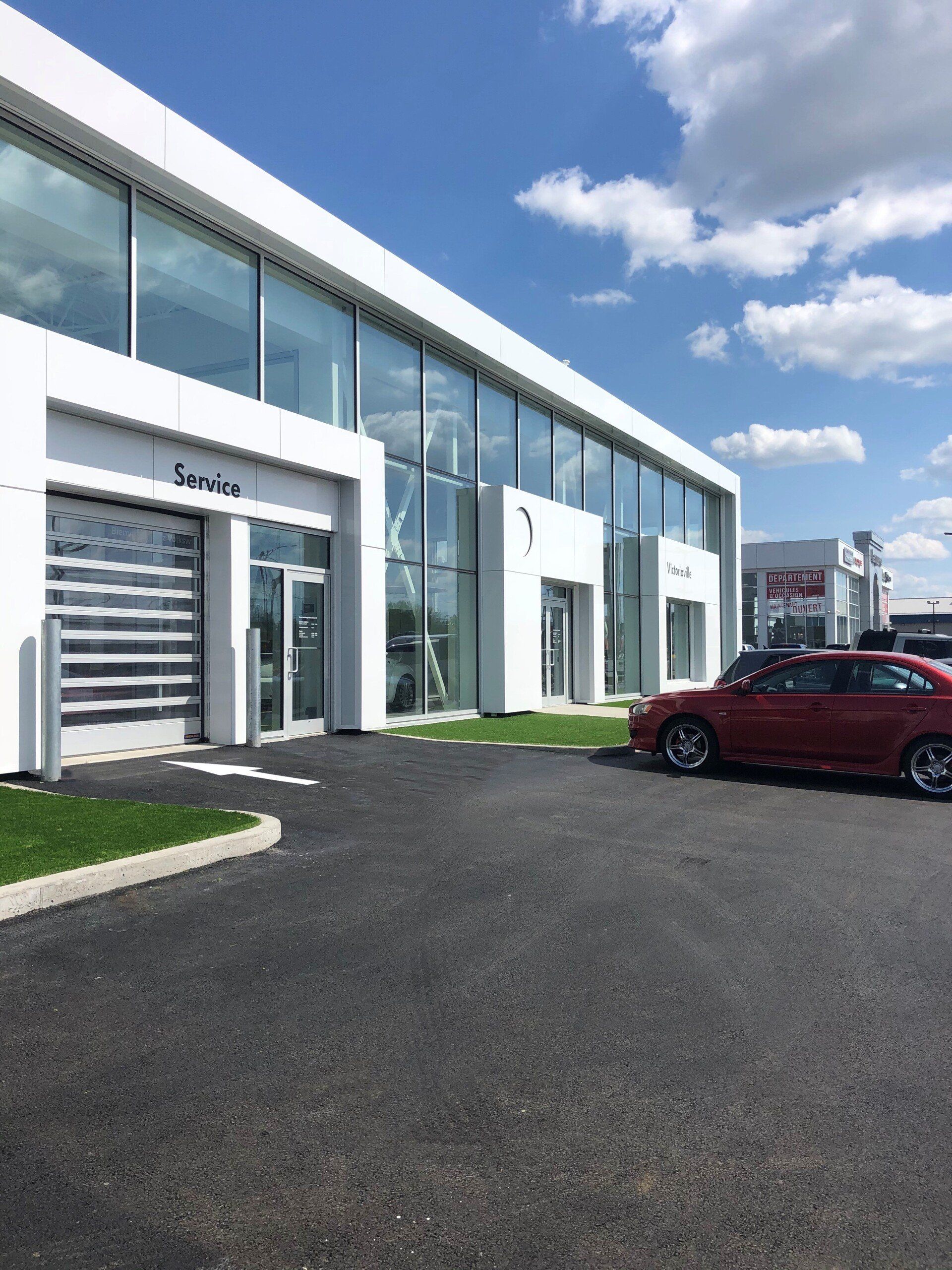 Bâtiment blanc moderne avec de grandes fenêtres et une aire de service. Voiture rouge garée devant. Journée ensoleillée.