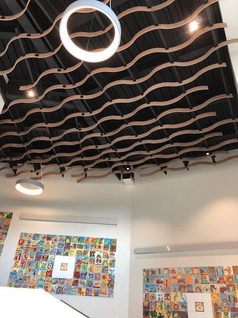 Plafond à lattes de bois ondulées et luminaires circulaires au-dessus de murs blancs ornés d'œuvres d'art colorées.