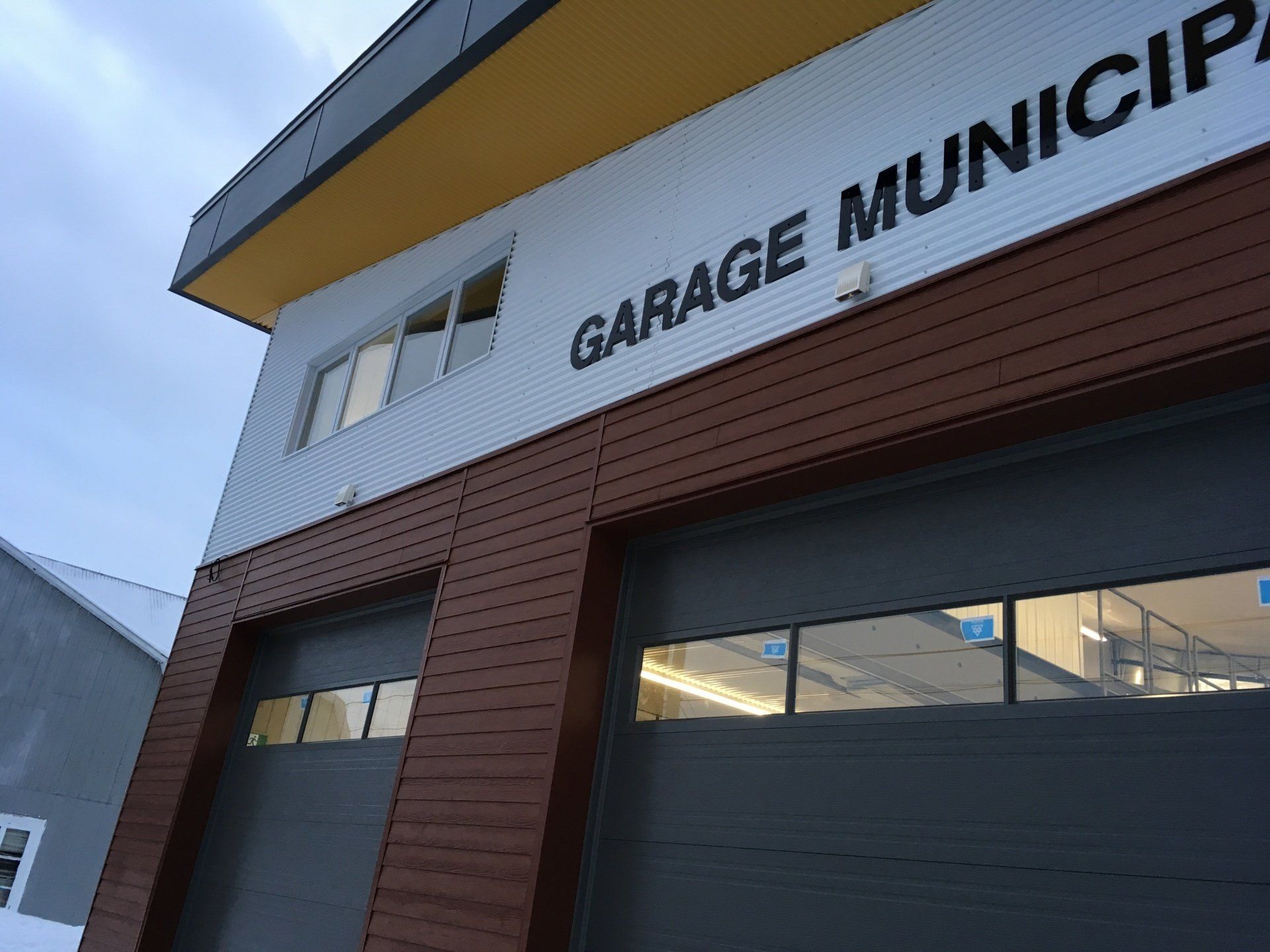 Bâtiment « Garage Municipal » avec portes de garage grises et accents bruns et jaunes, vue extérieure.