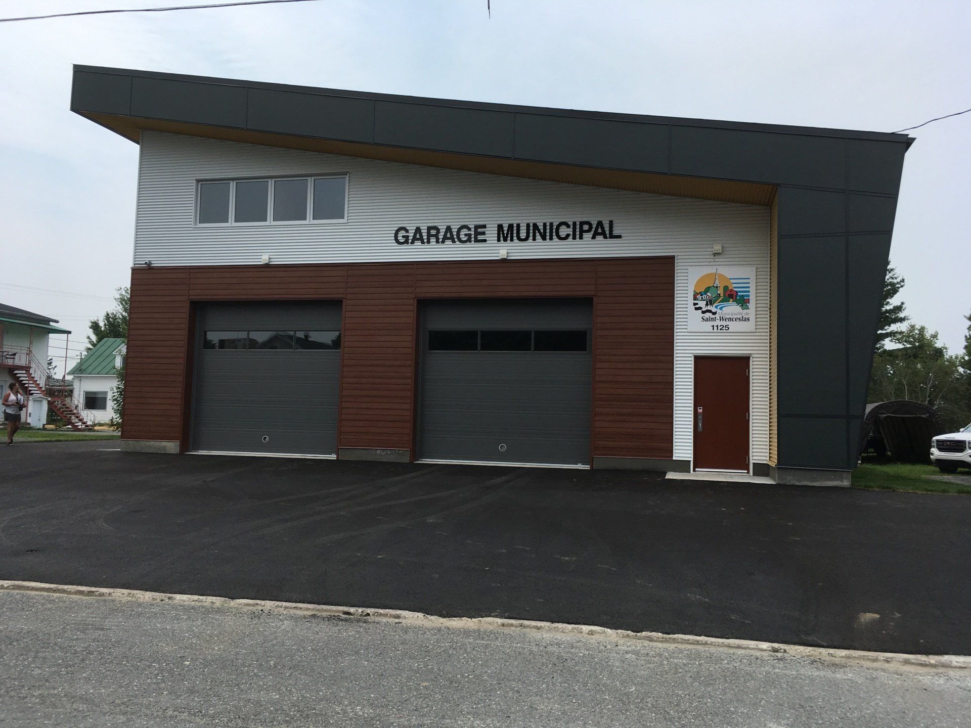 Bâtiment de garage municipal à deux travées, façade brune et grise, route asphaltée.