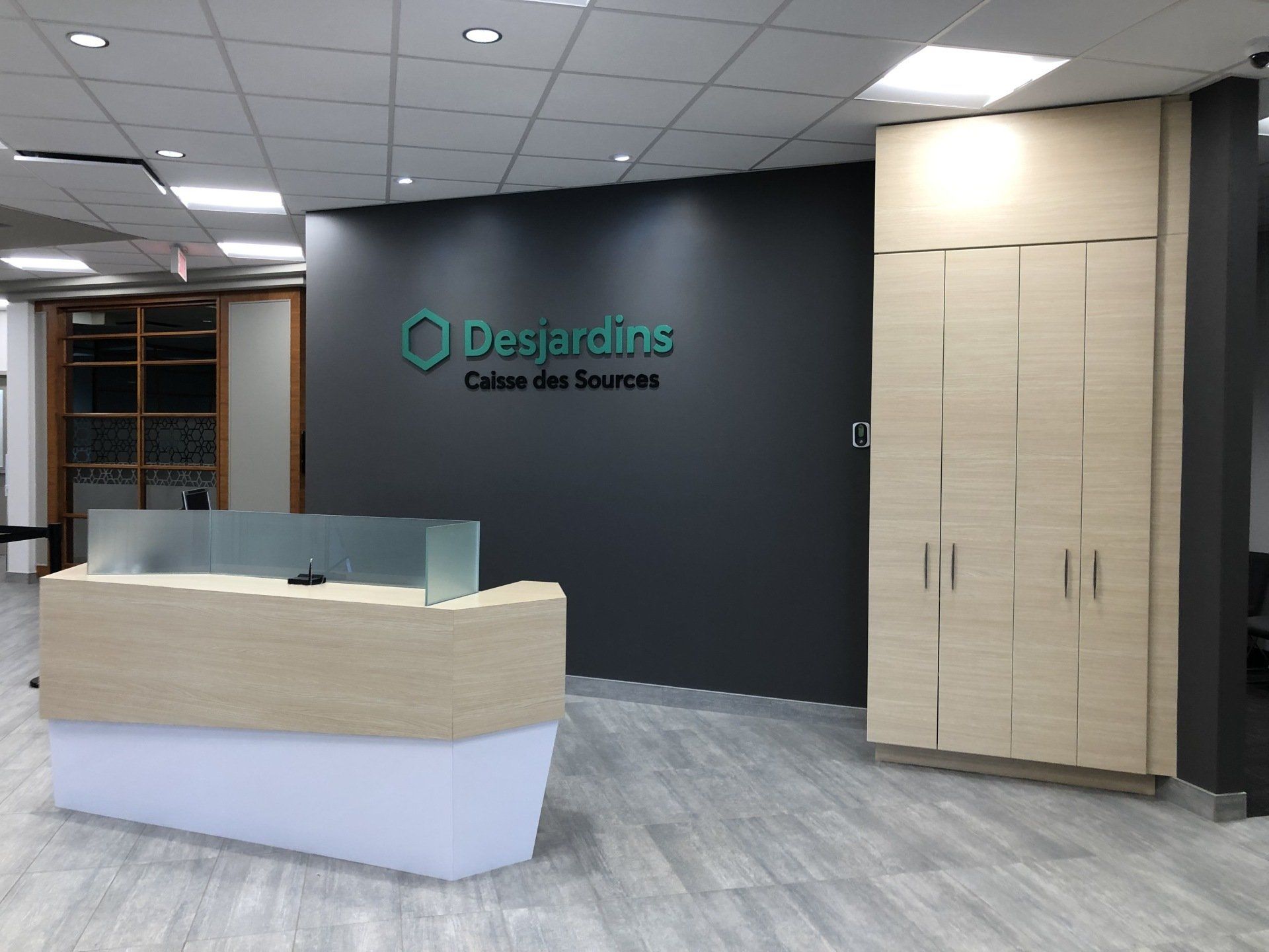 Entrée des bureaux de Desjardins avec comptoir d'accueil, logo sur un mur gris foncé et armoires en bois clair.