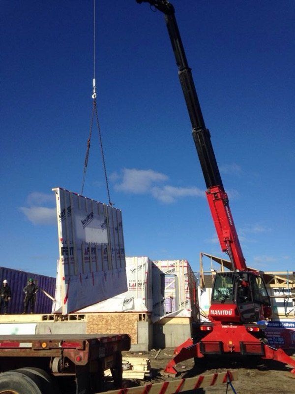 Une grue rouge soulève un grand panneau mural sur un chantier de construction, se détachant sur un ciel bleu.