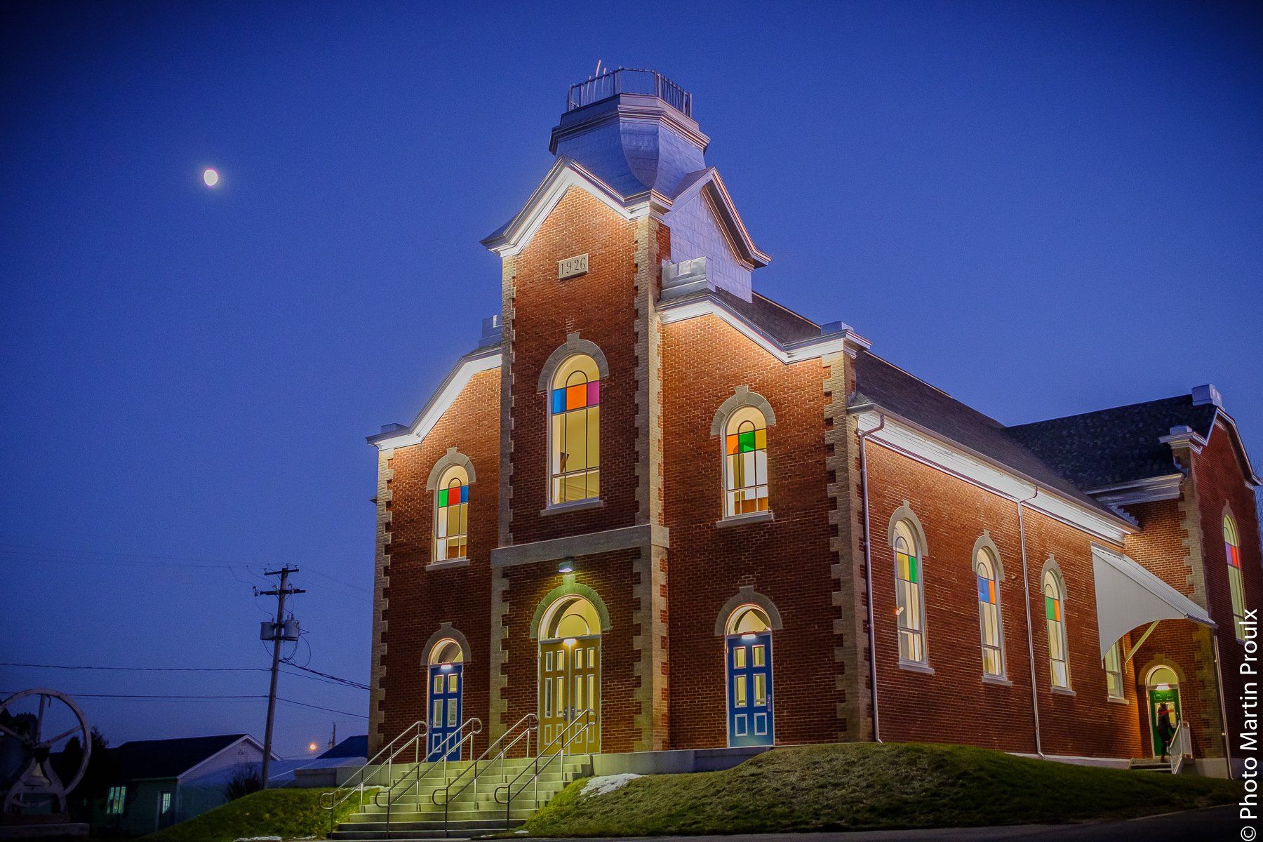 Église de briques au crépuscule, avec ses vitraux éclairés et la lune dans le ciel.