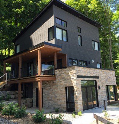 Maison moderne de trois étages avec bardage gris foncé, soubassement en pierre et terrasse en bois entourée d'arbres.