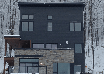 Maison noire moderne avec soubassement en pierre et grandes fenêtres, paysage enneigé.