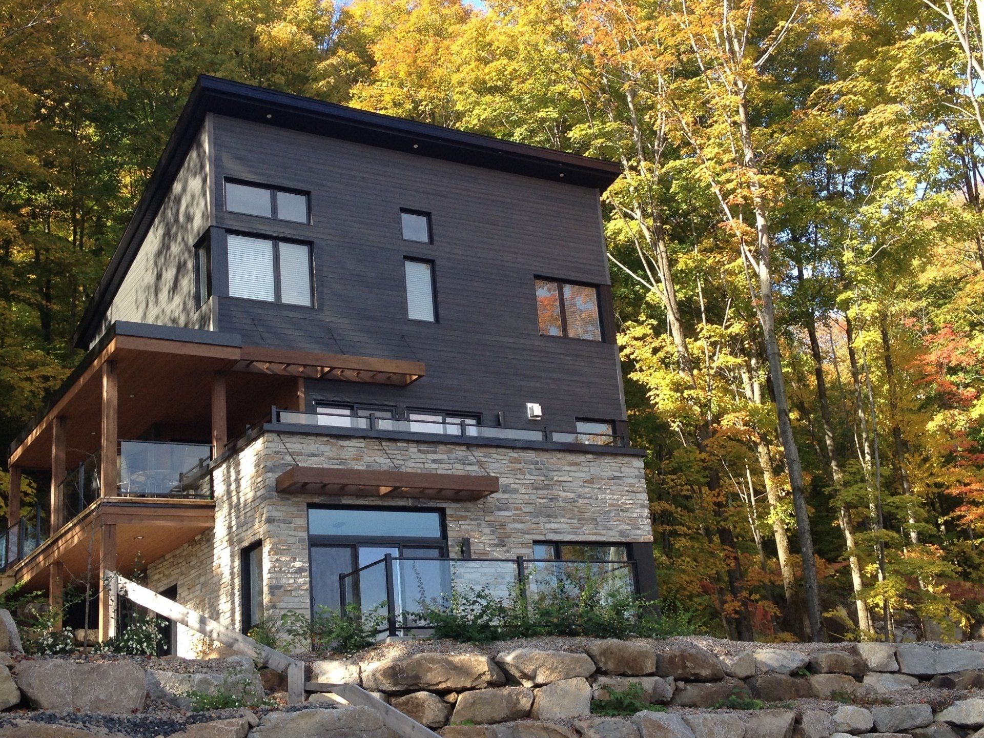 Maison moderne avec façade en pierre et bardage foncé, terrasse et arbres d'automne.