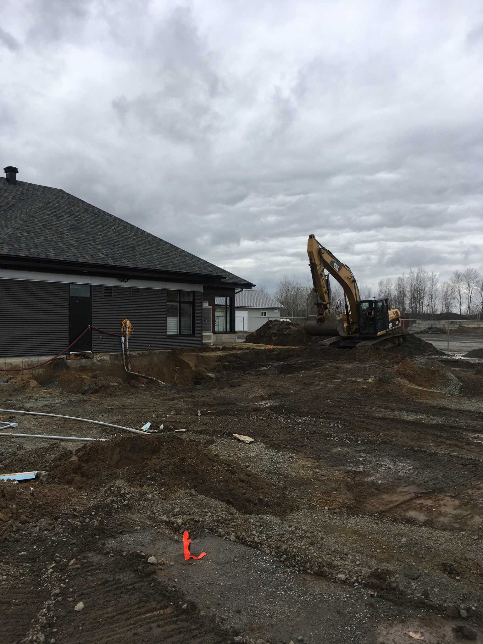 Chantier : Une pelleteuse se trouve près d'un bâtiment au revêtement sombre, sous un ciel nuageux.