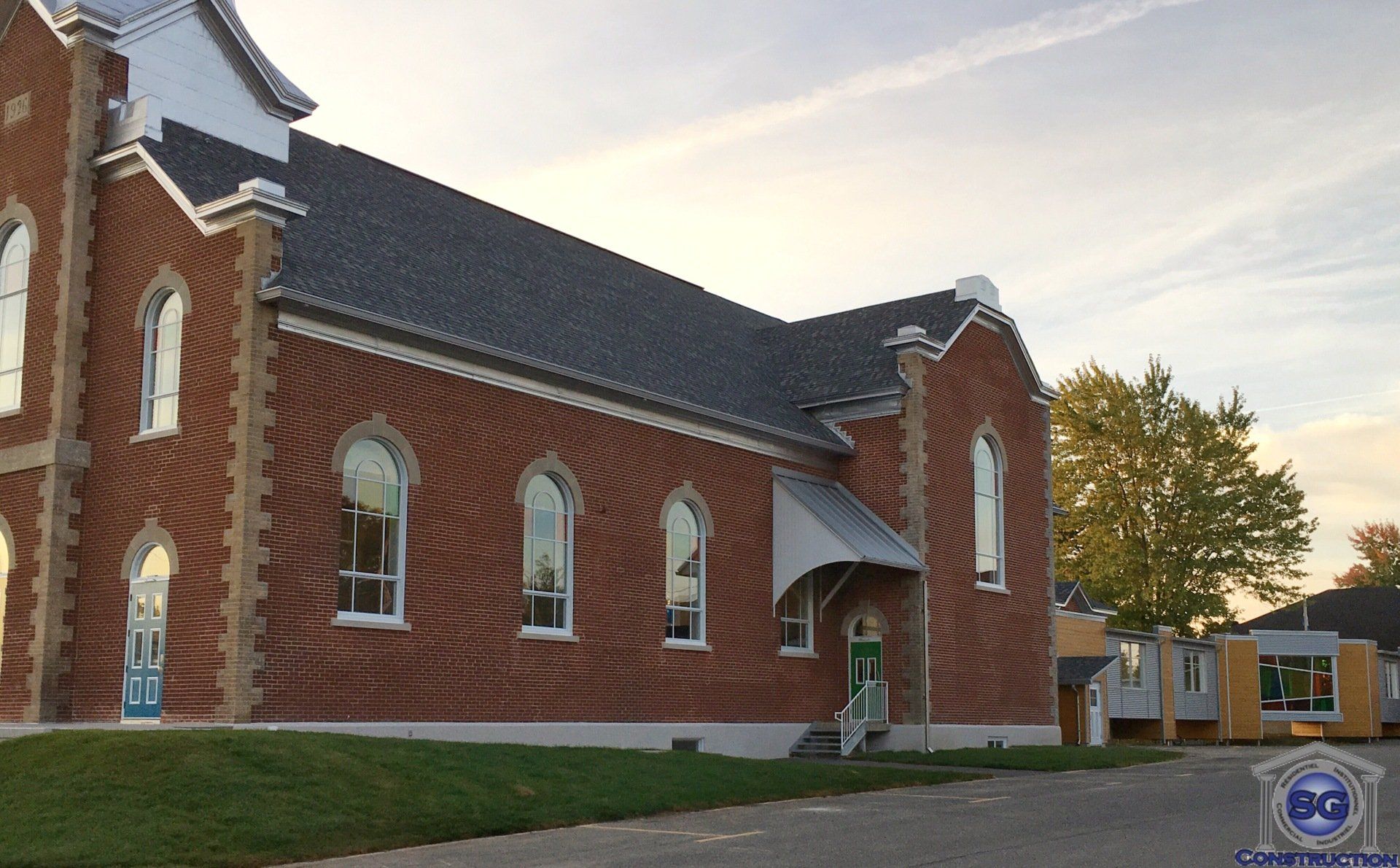 Église en briques rouges, toit gris et fenêtres cintrées. Porte verte avec auvent.