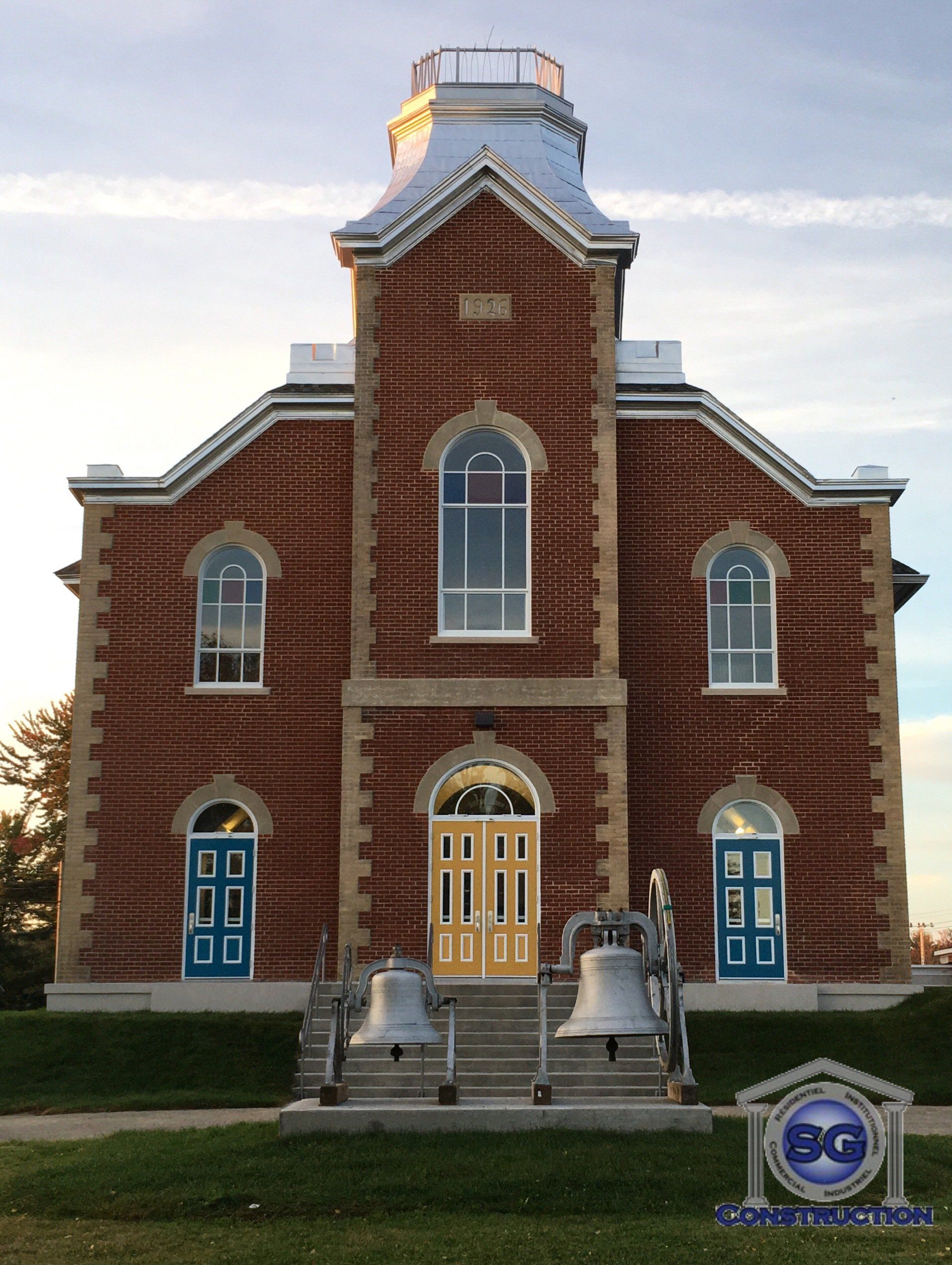 Bâtiment en briques avec tour, portes bleues et jaunes, et deux cloches au sol.