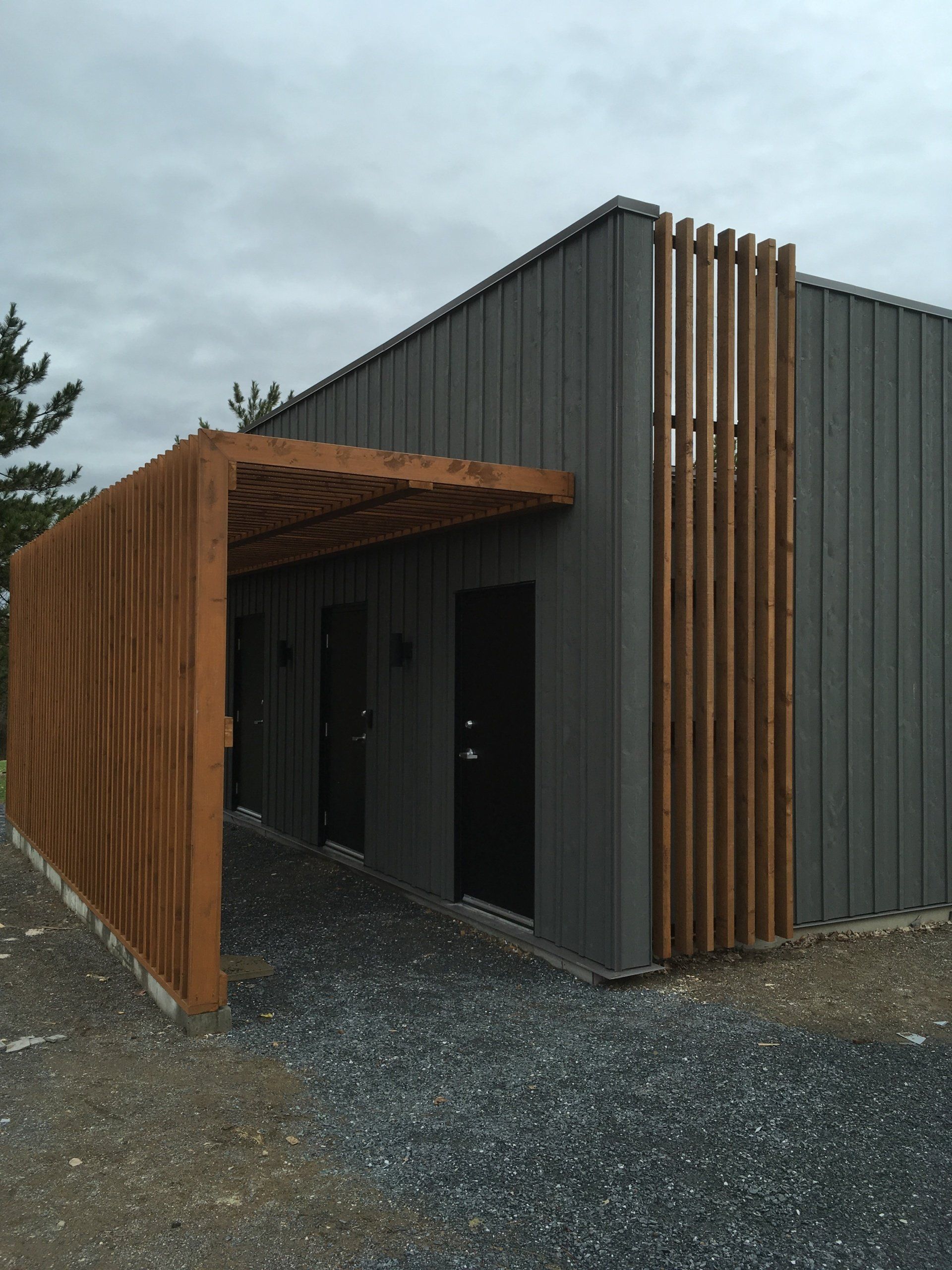 Bâtiment gris avec des éléments en bois, notamment une allée couverte et des lattes verticales. Sol en gravier.