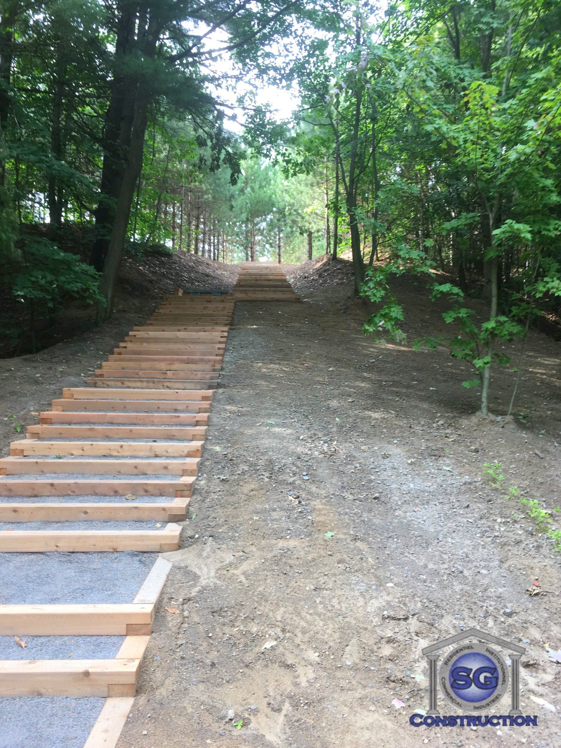 Escalier en bois aménagé à flanc de colline boisée. Chemin de gravier à côté.