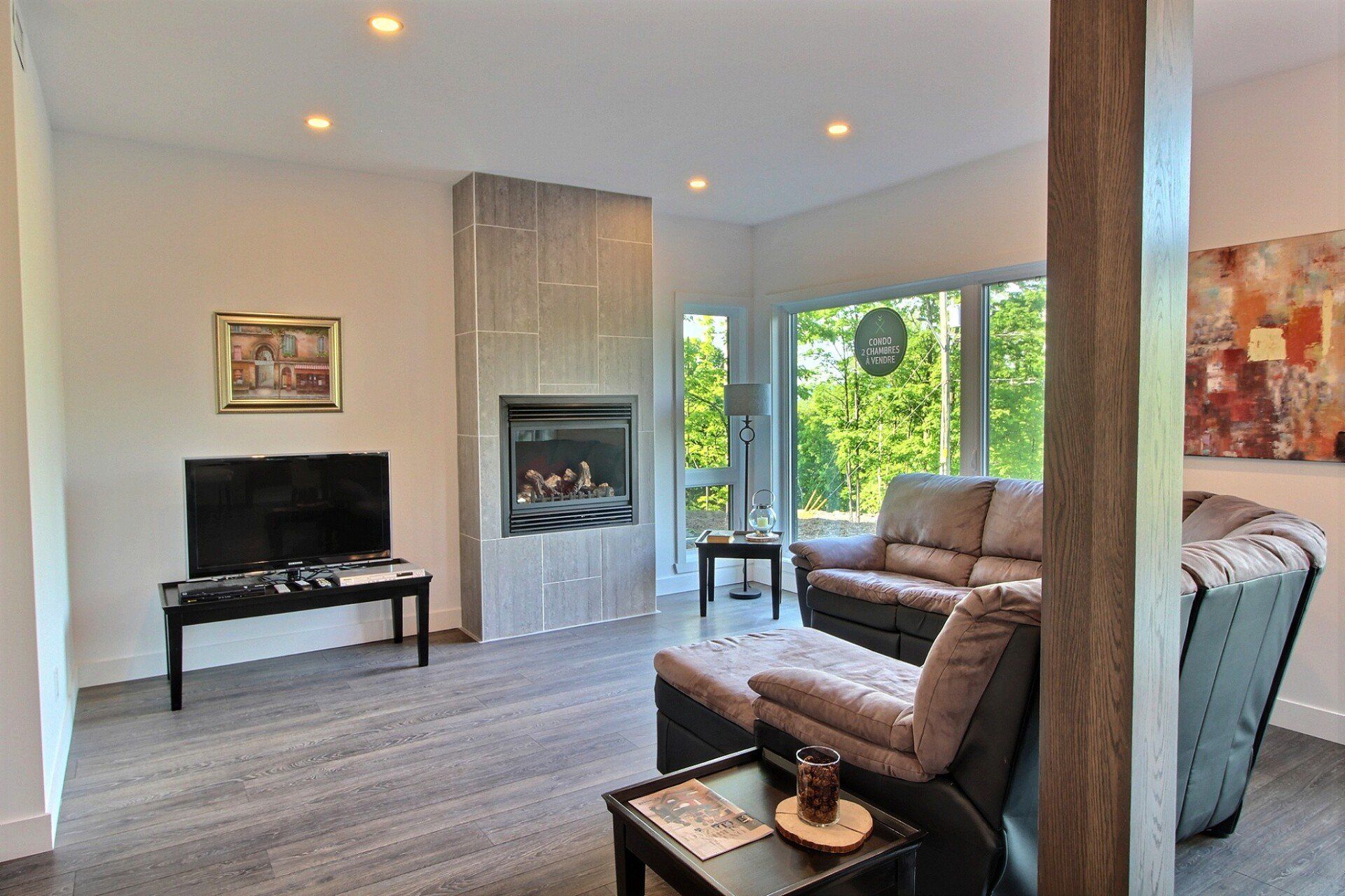 Salon avec sol gris, cheminée, canapé et grandes fenêtres donnant sur les arbres.