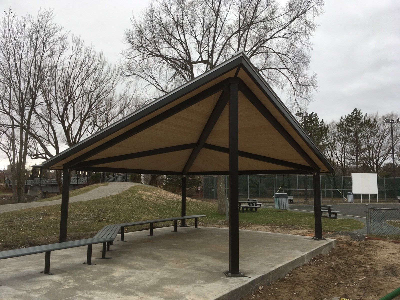 Abri à structure noire, toit beige et bancs dans un parc par temps nuageux.