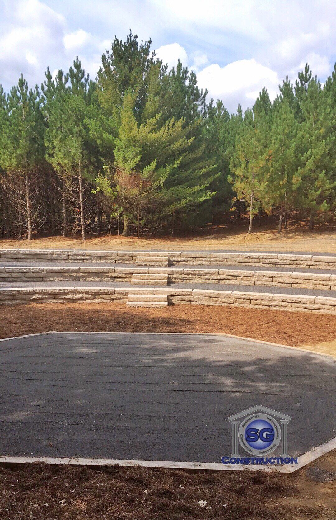 Un espace extérieur aménagé en gradins avec la forêt en arrière-plan. Le sol est sombre et les sièges sont en bois clair.