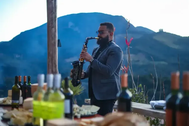 Músico tocando saxofone ao ar livre com montanhas ao fundo e garrafas sobre uma mesa em primeiro plano.