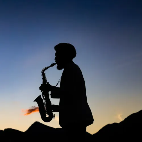 Silhueta de um saxofonista tocando ao ar livre ao entardecer contra um céu azul-alaranjado.